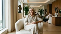 Mature woman with blonde hair, Caucasian ethnicity, wearing beige linen clothing, seated in cream upholstered armchair in modern luxury interior with wooden accents, potted plants, and natural light from large windows.