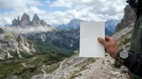 Hiker holding blank white card with alpine mountain peaks and valley landscape in background, showcasing mockup space for travel, adventure, and outdoor marketing campaigns.