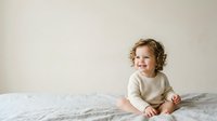 Toddler with curly blonde hair, Caucasian, wearing beige long-sleeve shirt, sitting on white bedding and smiling while looking away. Suitable for parenting, childcare, and family lifestyle content.