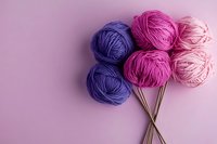 Colorful yarn balls arranged as flowers with knitting needles on pink background, featuring purple and pink wool skeins in creative composition.