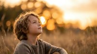 Young boy with curly hair sitting peacefully in golden sunset light, looking up with wonder and contemplation in natural outdoor setting.