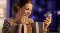 Young Caucasian woman smiling while using smartphone after shopping, holding paper bags in evening city lights. Online shopping and retail therapy concept.