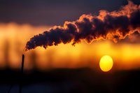 Industrial chimney emitting dark smoke against dramatic orange sunset sky with large glowing sun, highlighting environmental and climate change concerns for sustainability topics.