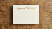 Elegant cream birthday card with gold script lettering on rustic wooden background for celebration greetings and party invitations.