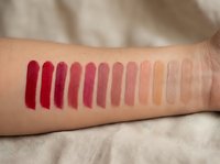 Lipstick color swatches on woman's arm showing gradient from deep red to nude shades for cosmetic testing and beauty product selection.