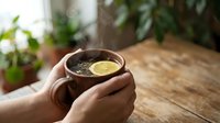 Hands holding warm herbal tea with lemon slice in ceramic mug on wooden table with blurred green plants in background, suitable for wellness and lifestyle content.