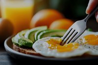 Sunny side up eggs on avocado toast with fork piercing runny yolk, served with fresh oranges. Close-up macro shot of healthy breakfast on rustic wooden table.