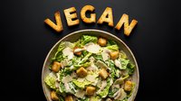 Vegan Caesar salad in a ceramic bowl with croutons and parmesan shavings, topped with croutons spelling VEGAN on dark background.