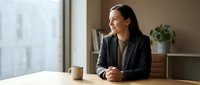 Professional Asian businesswoman in dark suit sitting at wooden desk near window, contemplating future plans in modern office environment.