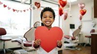 Happy African American boy holding red heart shape in decorated dental office during Valentine's Day celebration with festive balloons and bunting.