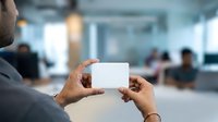 Professional holding blank white card in modern office environment with colleagues working in background, suitable for business branding and corporate communications.