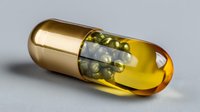 Golden pharmaceutical capsule with transparent amber end revealing medicine beads inside, photographed on neutral gray background for healthcare and medical concepts.