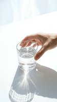 Hand reaching for a clear glass of fresh water on a white surface with sunlight casting a bright shadow pattern, symbolizing hydration and wellness.