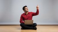Young South Asian male in red shirt and dark pants sitting cross-legged on wooden floor with laptop, pointing upward with confident expression against neutral background for business and technology concepts.