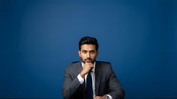 Confident male professional with dark hair and beard wearing gray suit and tie, hand near chin in thoughtful pose against solid blue background for corporate and business concepts.