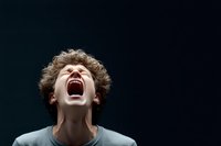 Young Caucasian male with curly hair screaming in intense emotional expression against dark background, head tilted back showing raw feelings and vulnerability.