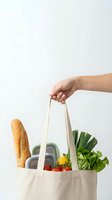 Hand holding eco-friendly canvas tote bag filled with fresh groceries including bread, vegetables and containers on white background for sustainable shopping.