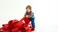 Young toddler boy in striped shirt and overalls playing with large red ribbon bow on white background, holiday gift concept for seasonal marketing.