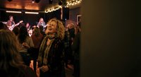 Joyful blonde woman dancing and laughing at live music concert with band performing on stage in intimate venue with warm lighting and enthusiastic crowd.