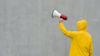 Person in bright yellow hoodie holding megaphone against concrete wall background for protest communication and activism campaigns.
