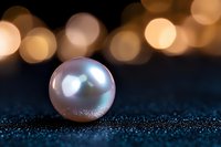Single luminous pearl on dark textured surface with golden bokeh lights in background creating magical atmospheric composition for luxury design.