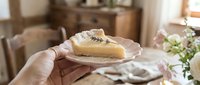 Hand holding slice of homemade lemon tart with meringue topping on white plate in cozy kitchen setting with rustic wooden furniture and fresh flowers.