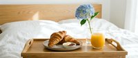 Breakfast in bed with fresh croissants, orange juice and blue hydrangea flower on wooden tray over white bedding for romantic morning meal service.