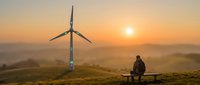 Person sitting on bench watching sunrise with wind turbine on hill generating clean renewable energy in peaceful countryside landscape.