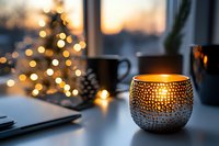 Cozy winter evening atmosphere with glowing candle holder and blurred Christmas tree lights in background, creating warm holiday ambiance at home workspace.