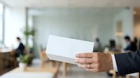 Close-up of businessman hand holding blank white business card in modern office environment with blurred colleagues and workspace in background.