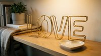 Gold wire LOVE letters decor on wooden shelf with cozy home interior, plant and blanket in background.
