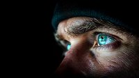 Intense close-up of glowing cyan eyes in dark setting shows male face partially hidden by beanie hat, creating mysterious futuristic cyberpunk atmosphere.