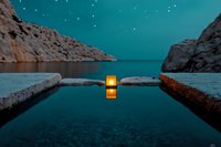 Glowing lantern on stone pier reflects in calm water between rocky cliffs under starry turquoise night sky, creating serene atmospheric scene.