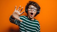 Excited young boy with curly hair wearing headphones and glasses gestures with outstretched hand against vibrant orange background, expressing surprise.