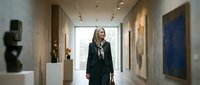 Woman with long dark hair, wearing a black blazer and patterned scarf, walking through a contemporary art gallery with sculptures and paintings displayed on white walls and pedestals.