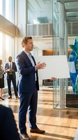 Confident businessman presenting blank poster in modern office lobby with colleagues in background for corporate communication and marketing campaigns.