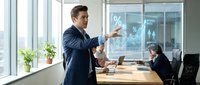 Caucasian male businessman in dark blue suit presenting financial data on glass board to female colleagues in modern office conference room with large windows and natural light.