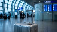 Silver suitcase in a modern airport terminal near arrival board, travelers in background, travel and tourism concept.