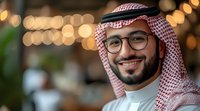 Young Middle Eastern man wearing traditional white thobe and red-checkered ghutra headwear with glasses, smiling at camera in modern interior setting with warm bokeh lighting.
