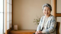Senior Asian woman with gray hair smiling at camera in bright home interior, wearing light cardigan over white top, seated by window with natural lighting for lifestyle and wellness content.
