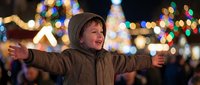 Young boy with arms outstretched enjoying festive holiday lights at evening Christmas market. Child expressing joy and freedom during winter celebration.