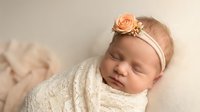 Peaceful newborn baby girl sleeping wrapped in cream knit blanket wearing peach flower headband for maternity photography and nursery decor.