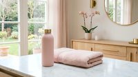 Spa wellness setup with folded pink towels, cosmetic bottle and orchid flowers on marble countertop near windows, home bathroom decor.