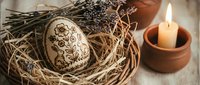 Decorative wooden Easter egg with intricate floral patterns nestled in natural straw nest beside burning candle and dried lavender sprigs on rustic surface.