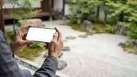 Man holding smartphone with blank white screen outdoors in garden setting, displaying mobile device for app mockup or website design presentation.