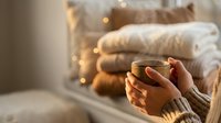 Cozy winter morning with hands holding warm coffee mug, soft blankets and fairy lights creating hygge atmosphere for relaxation and comfort at home.
