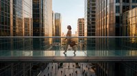 Professional businesswoman walking on modern glass bridge between skyscrapers during golden hour sunset in urban financial district downtown cityscape.