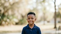 Young African American boy with short dark hair wearing a navy blue polo shirt, smiling at camera outdoors in park setting with blurred trees and natural daylight background.