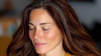 Young woman with closed eyes and serene expression, long brown hair flowing naturally, warm lighting creates peaceful ambiance against blurred background.