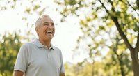 Senior man with gray hair and light skin tone, wearing a light gray polo shirt, smiling outdoors in a park setting with blurred green trees and natural sunlight, expressing joy and contentment.